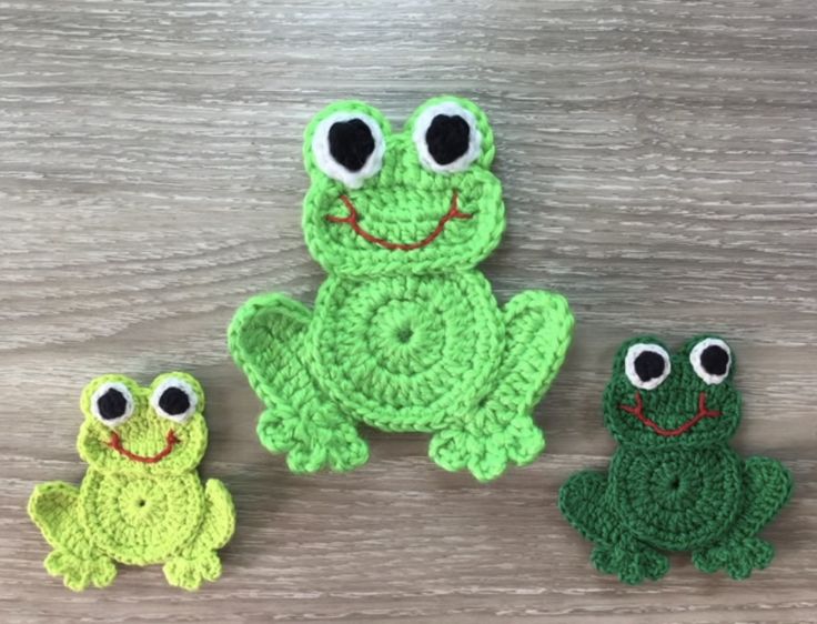 three crocheted frogs sitting next to each other on a wooden surface with eyes