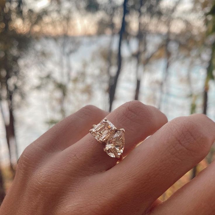 a woman's hand holding an engagement ring in front of some trees and water