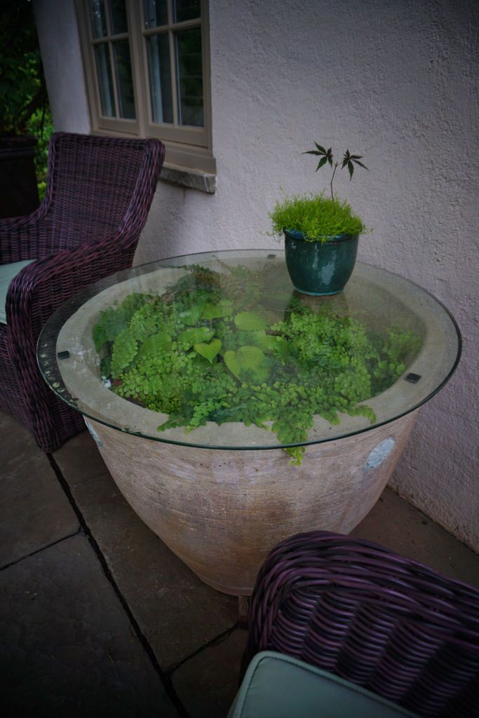 a glass table topped with green plants on top of a stone floor next to a purple wicker chair