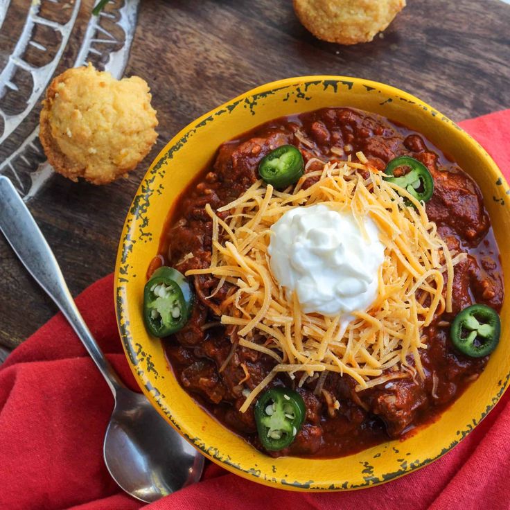 a yellow bowl filled with chili and cheese next to muffins on a wooden table