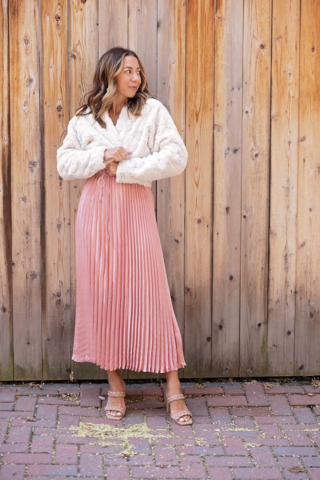 a woman standing in front of a wooden fence wearing a pink pleated skirt and white sweater