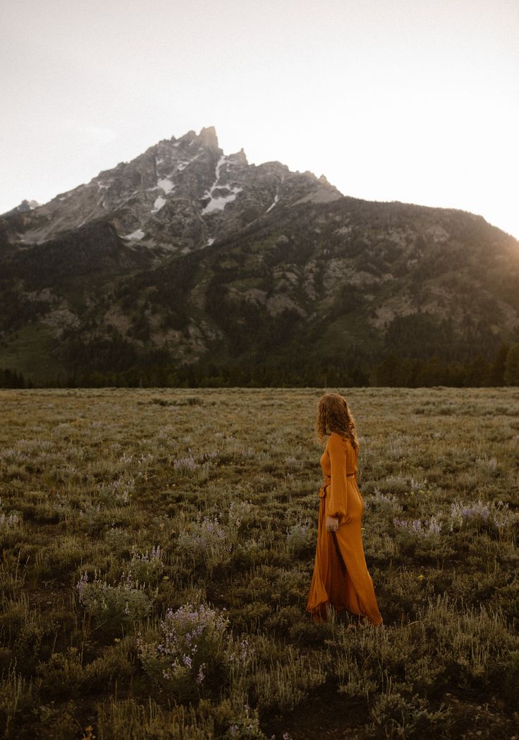 a woman in an orange dress standing in a field with mountains in the background at sunset