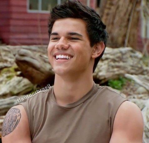 a young man with tattoos on his arm and chest smiles at the camera while sitting in front of a log cabin