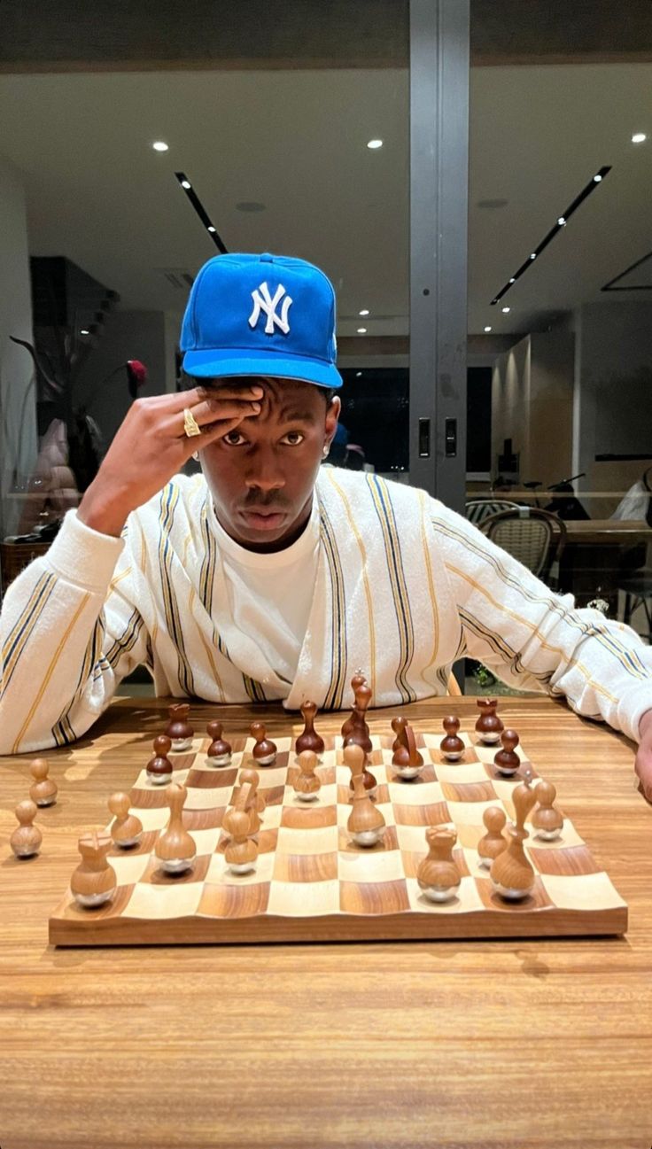 a young man sitting at a table with a chess board and hat on his head