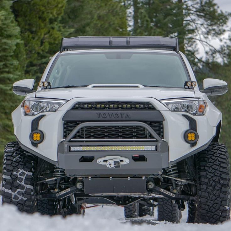 the front end of a white truck driving on snow covered ground with trees in the background