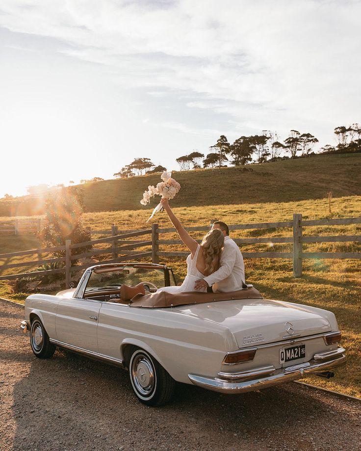 a man and woman sitting in the back of a white convertible car holding up flowers