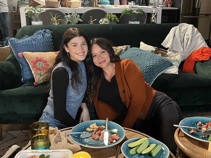 two women are sitting on a couch with plates of food in front of them,