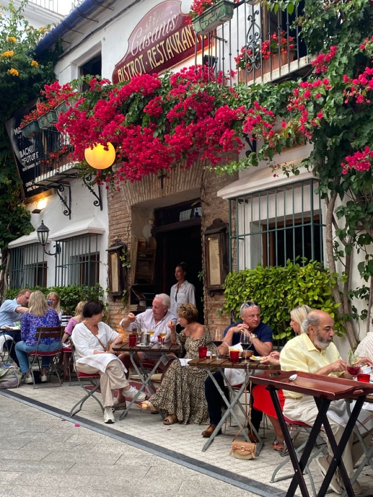 people are sitting at tables outside in front of a building with red flowers on it