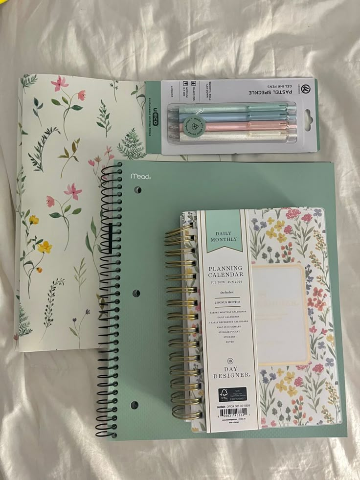 two notebooks and a pen sitting on top of a white sheet with floral designs