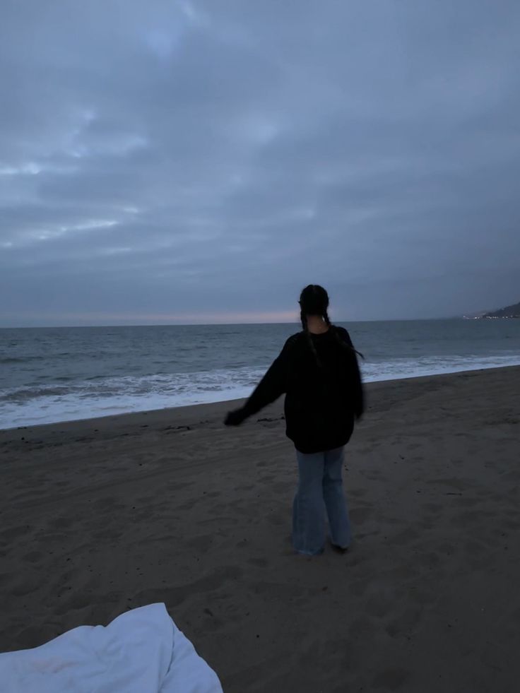 a woman standing on top of a sandy beach next to the ocean at dusk with her arms outstretched