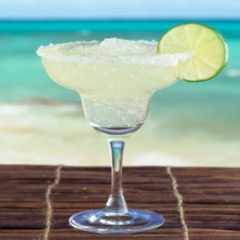 a margarita sitting on top of a wooden table next to the ocean with a lime wedge in it