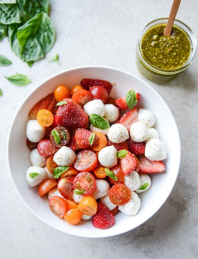 a white bowl filled with strawberries, mozzarella and basil next to a jar of pesto