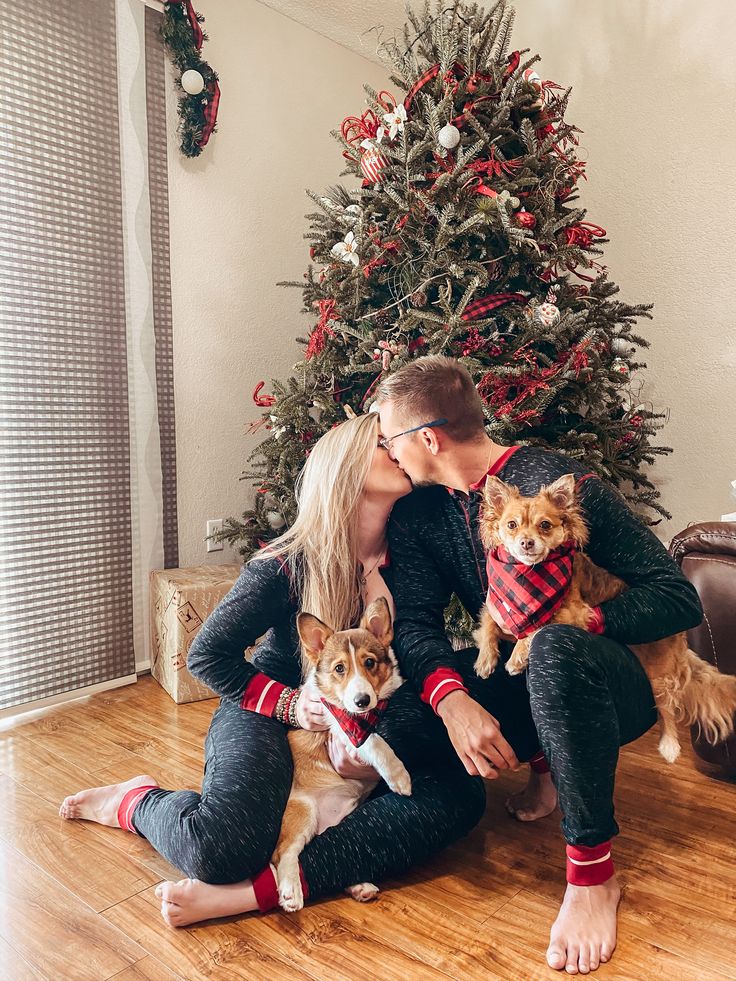 a man and woman kissing in front of a christmas tree with two dogs on their lap