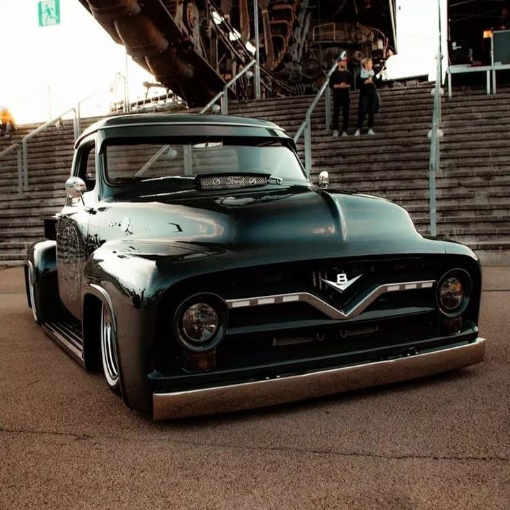 an old black truck parked in front of some bleachers with people walking up the stairs