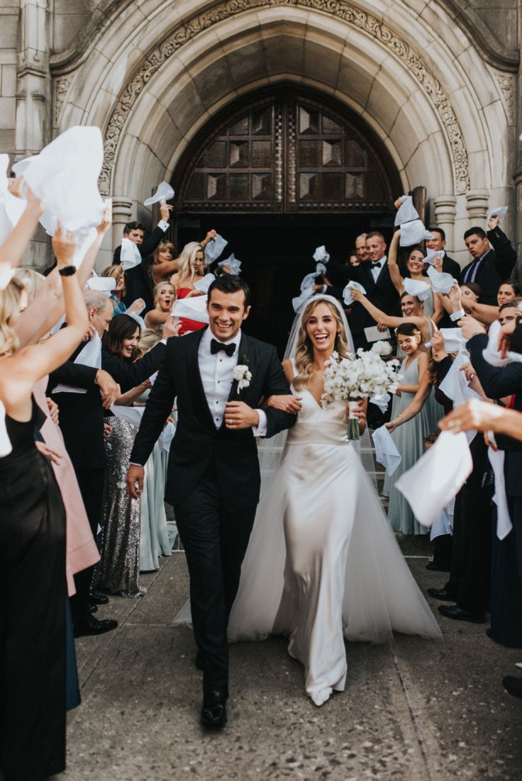 a bride and groom walk through the crowd as they exit an old building with confetti in their hands