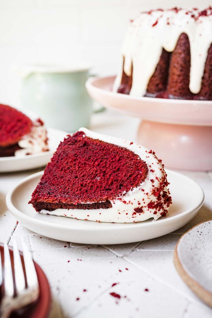 a slice of red velvet cake with white frosting on two plates next to the rest of the cake