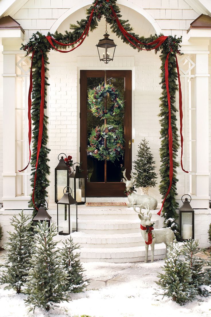 a white house decorated for christmas with wreaths and garland on the front door, lanterns and trees