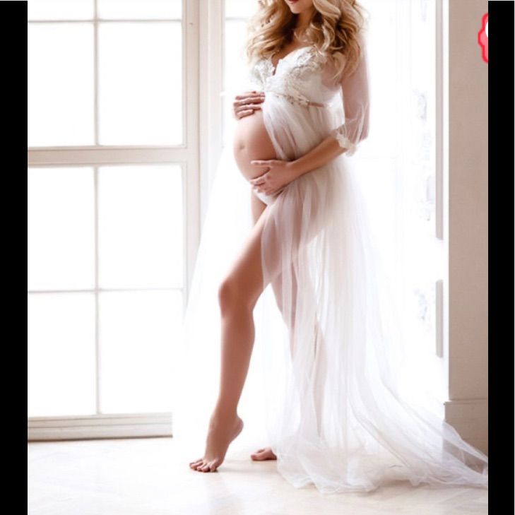 a pregnant woman is posing in front of a window wearing a white gown and holding her belly