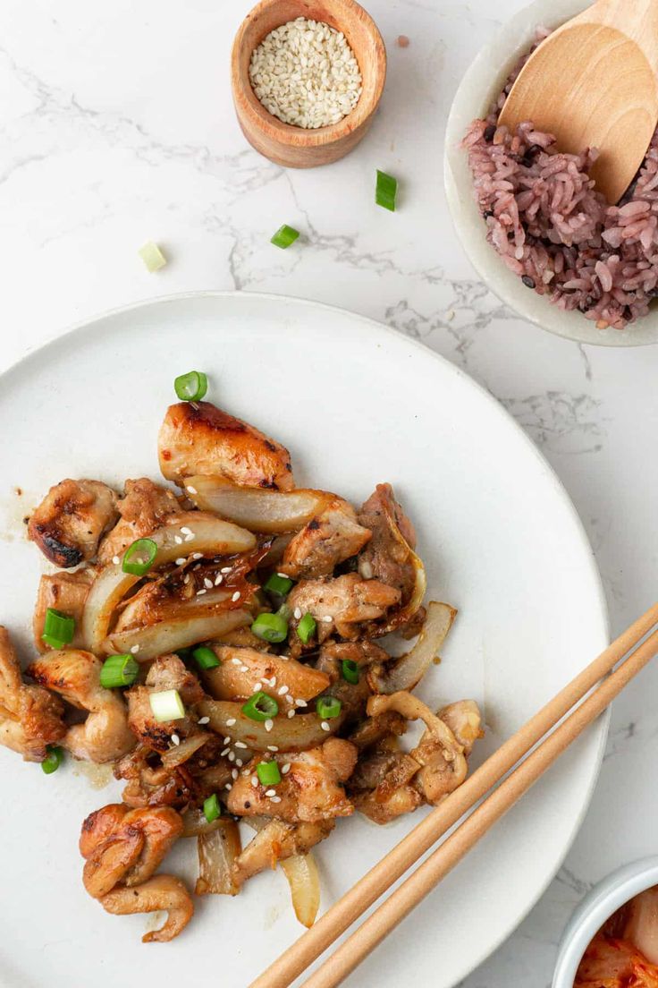 chicken and onions on a white plate with chopsticks next to the bowl full of rice