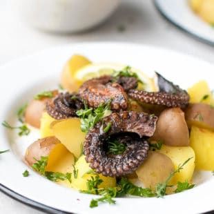 an octopus and potatoes dish on a white plate with lemon wedges, parsley