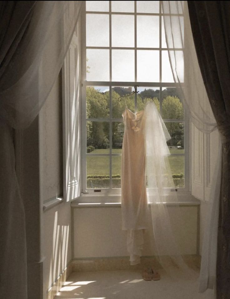 a bride's wedding dress hangs in front of a window with sheer drapes