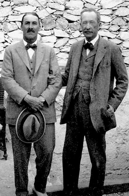 two men standing next to each other in front of a stone wall wearing suits and hats