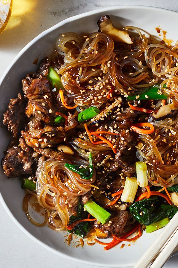 a white plate topped with meat and veggies covered in sesame seeds next to chopsticks