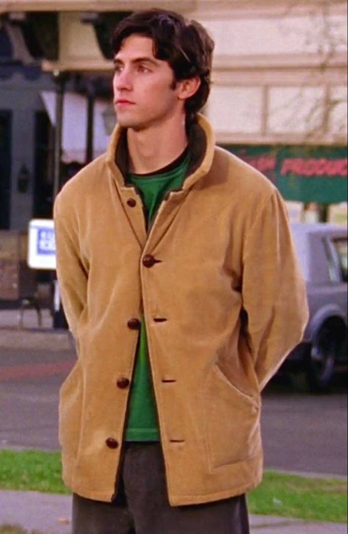 a young man standing in front of a building wearing a brown jacket and green shirt