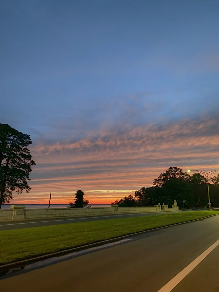 the sun is setting on an empty road with trees in the foreground and clouds in the background