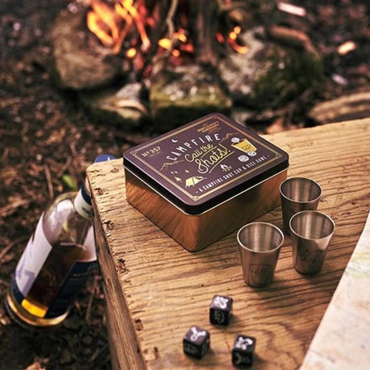 a wooden table topped with metal cups next to a campfire filled with wood burning logs