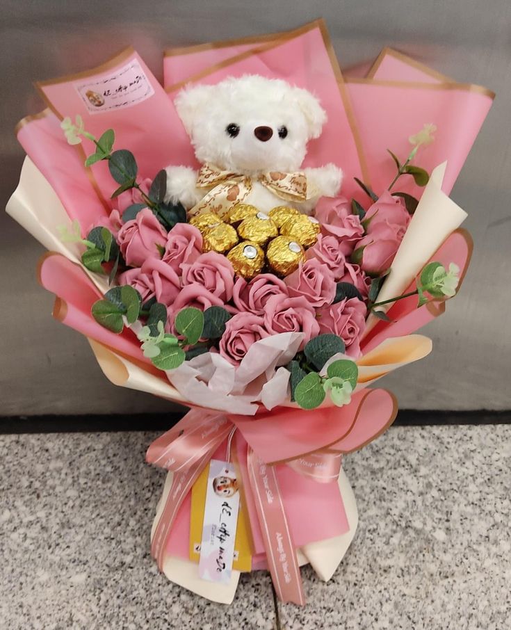 a teddy bear sitting on top of a bouquet of pink roses and chocolates in front of a wall