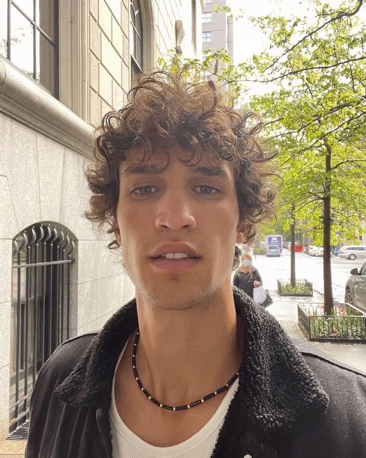 a young man with curly hair standing in front of a building on a city street