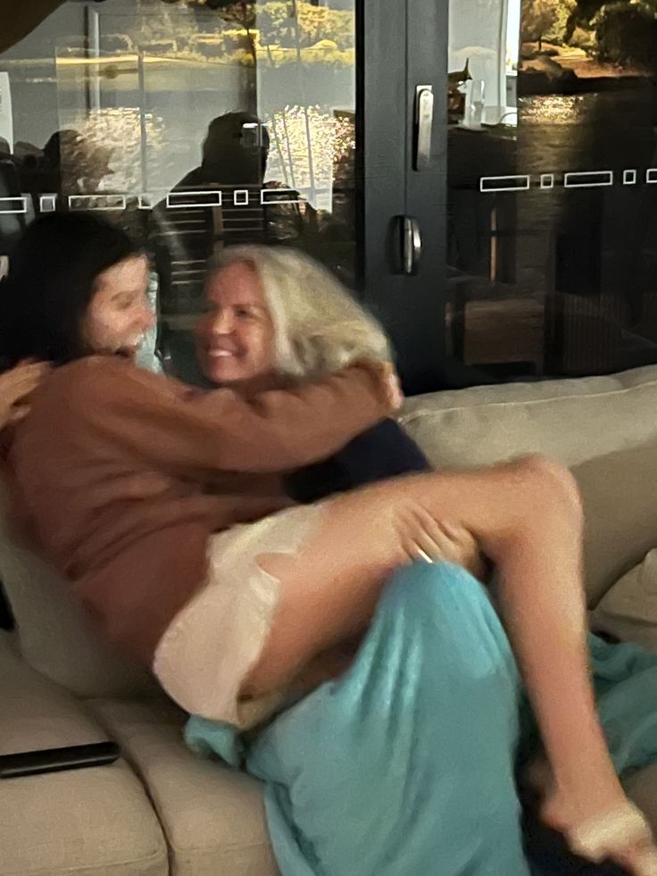 two women hugging each other while sitting on a couch in front of a glass window