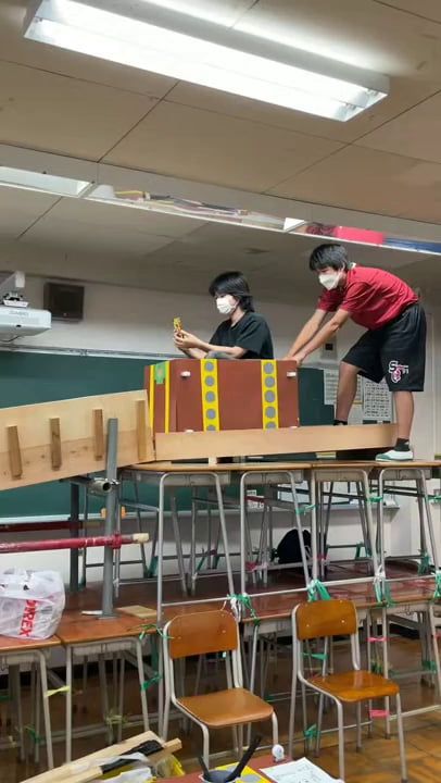 two boys wearing face masks are playing with wooden train tracks in a classroom filled with desks and chairs