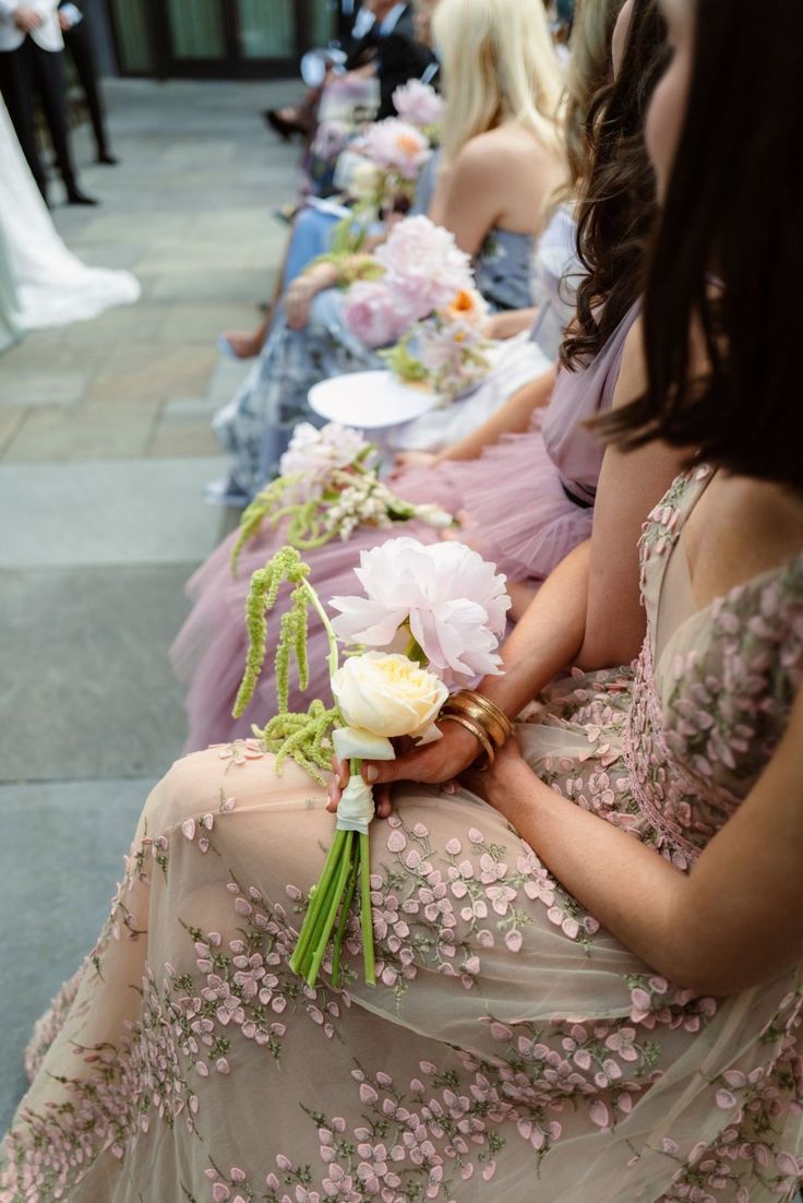 a row of bridesmaids in dresses sitting on the sidewalk with their bouquets