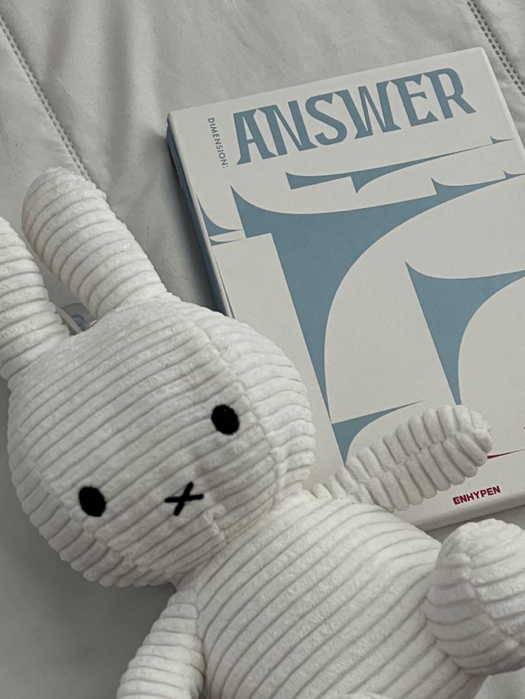 a white stuffed animal sitting next to an answer book on a bed with the cover pulled down