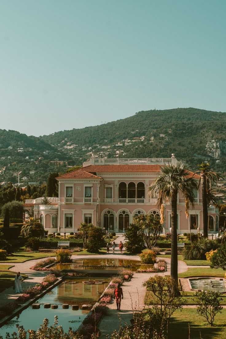 a large pink building surrounded by palm trees and greenery in front of a mountain