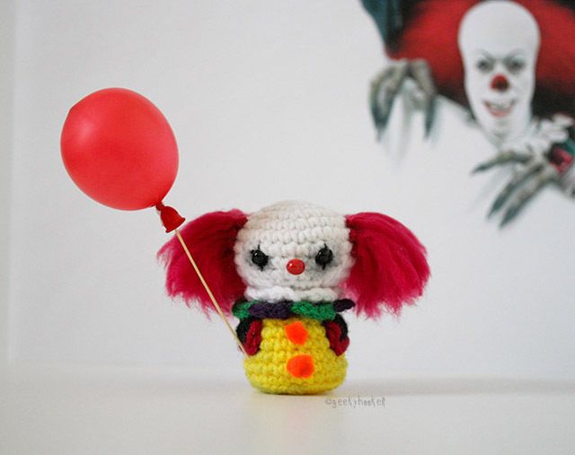 a crocheted clown holding a red balloon in front of a wall with a painting on it