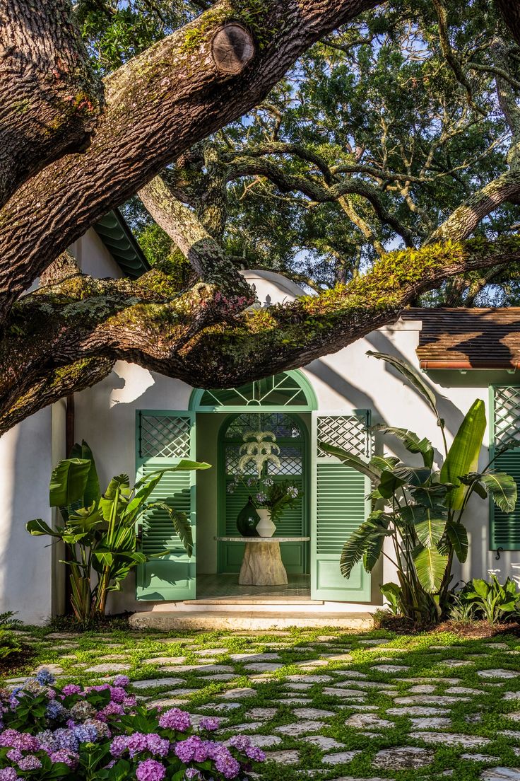 a house with green shutters and flowers in the front yard, surrounded by trees