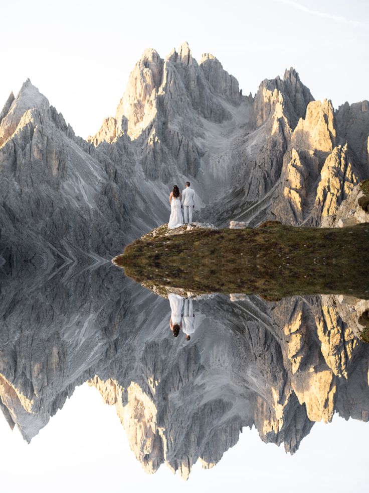 two people standing on top of a mountain next to a body of water with mountains in the background