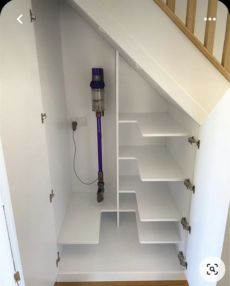 a white closet with shelves and a purple vacuum in the bottom half, under a stair case