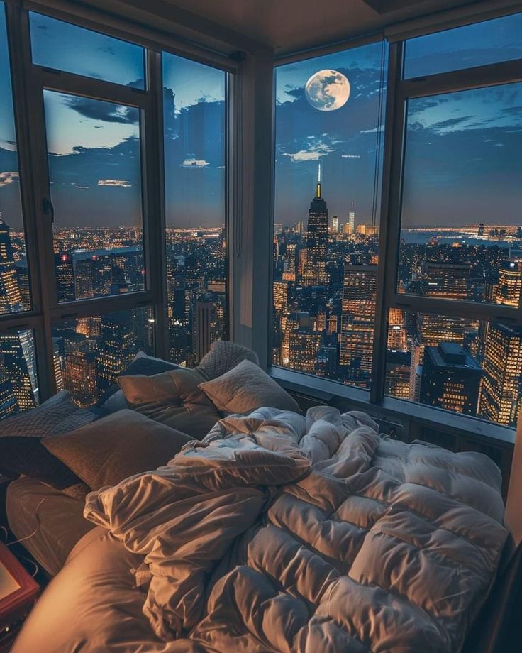 an unmade bed is in front of large windows overlooking the city at night time