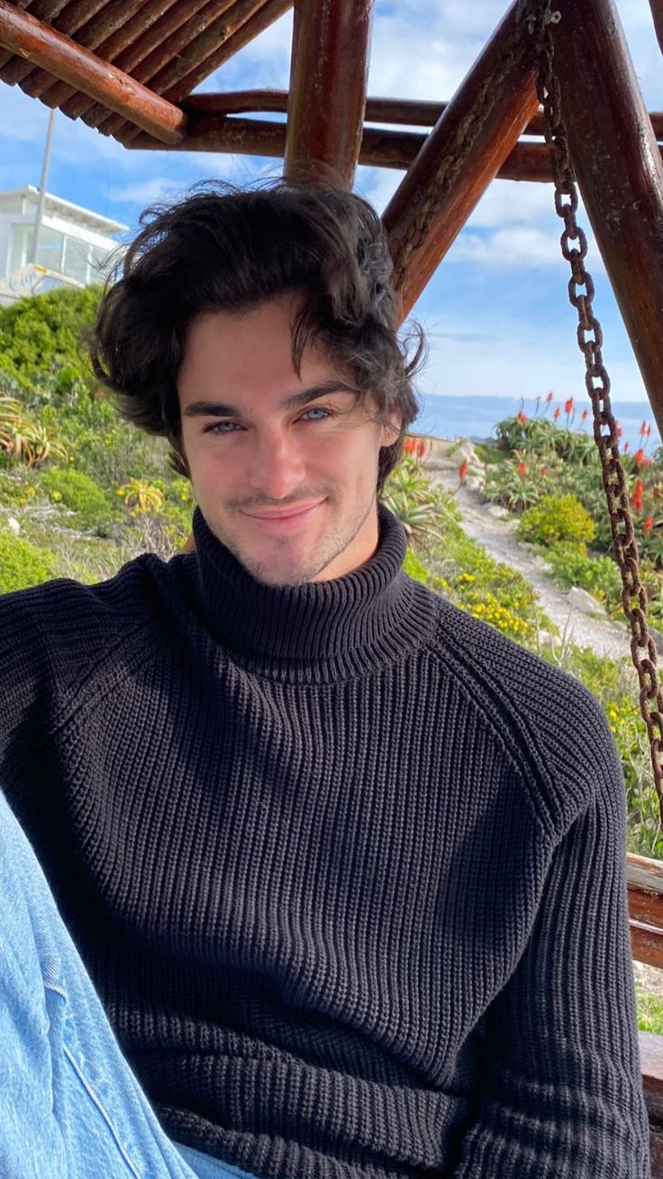 a man is sitting on a swing smiling at the camera while wearing a turtle neck sweater