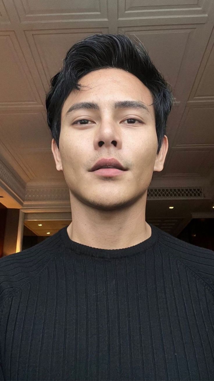 a young man with black hair and a sweater looking at the camera while standing in front of a ceiling