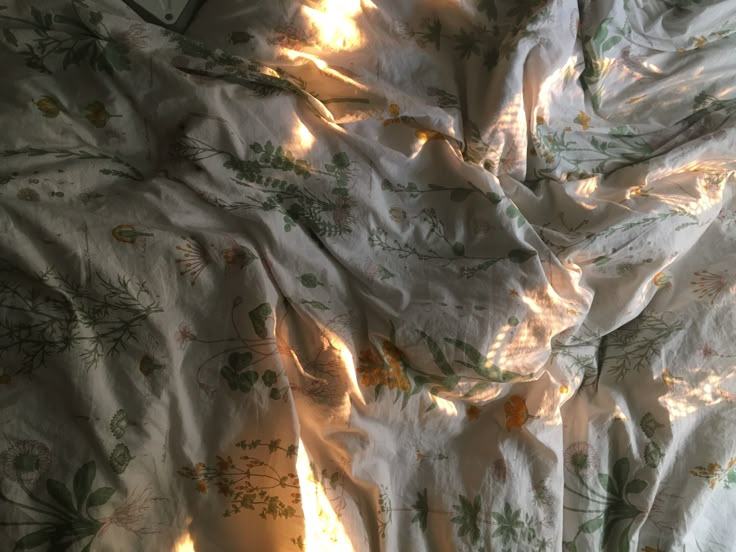 an unmade bed covered in white sheets with flowers on them and sunlight coming through the window