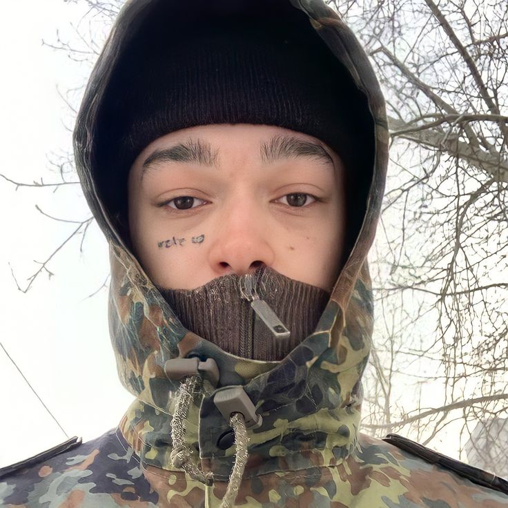 a young man wearing a camouflage jacket with a hoodie on his face and nose