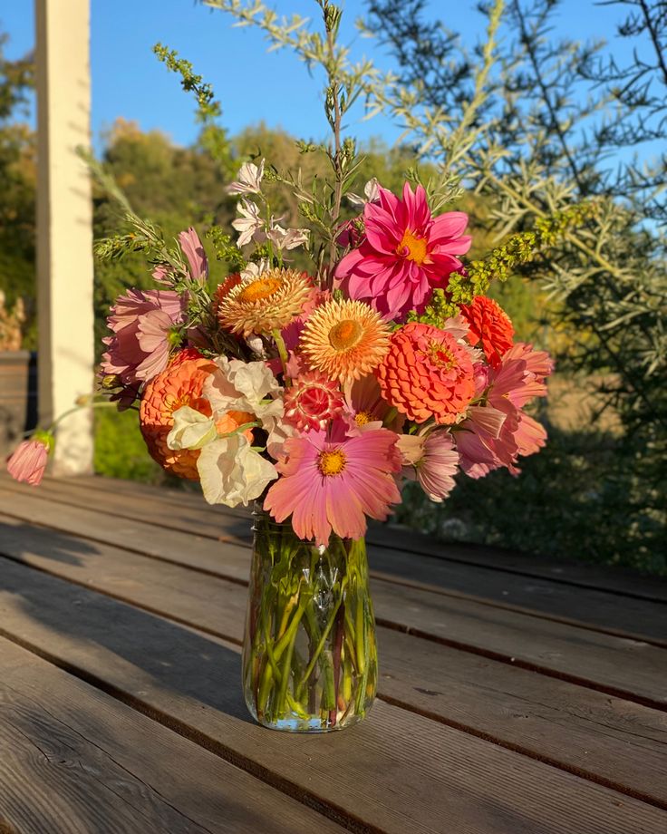 a vase filled with lots of different colored flowers on top of a wooden table next to trees