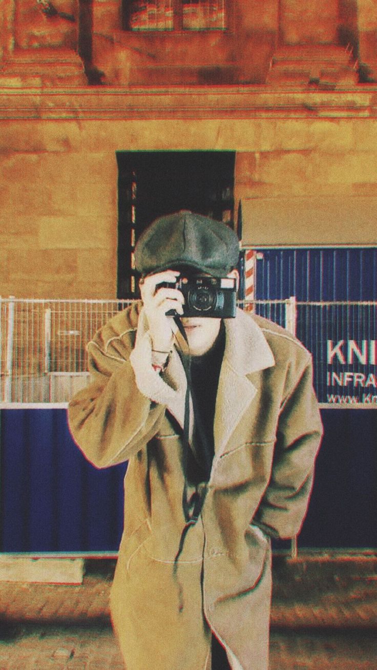 a man in a trench coat talking on a cell phone while holding a camera up to his face