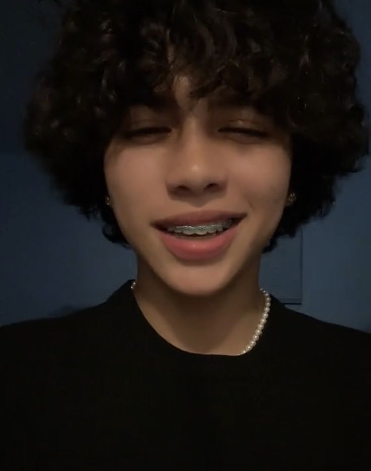 a young man with curly hair wearing a black shirt and pearls around his neck is smiling at the camera