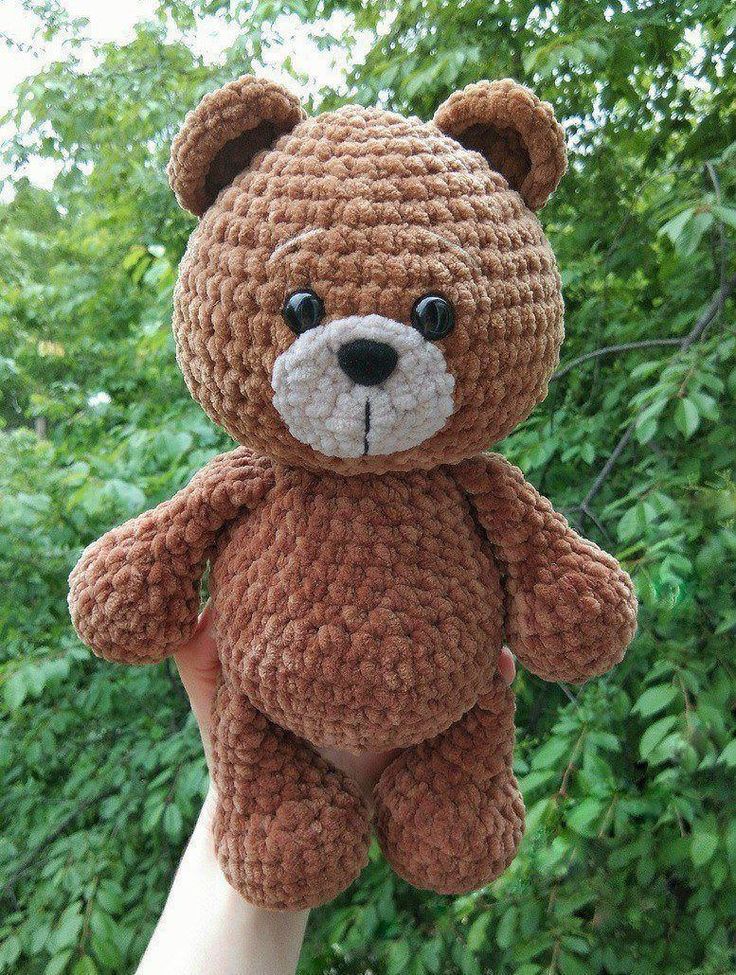 a hand holding a brown crocheted teddy bear in front of some trees and bushes